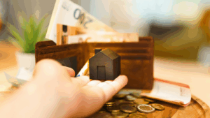 Person holding miniature home in front of loaded wallet and pile of change