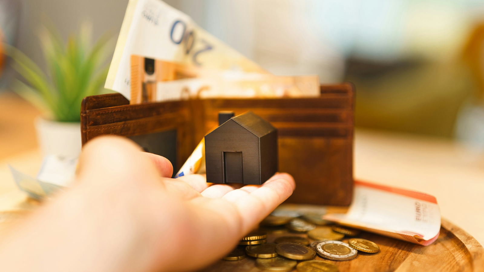 Person holding miniature home in front of loaded wallet and pile of change